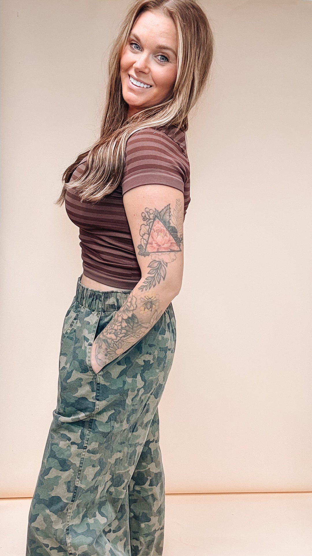 A woman with long blonde hair and arm tattoos is shown from the waist up, wearing a brown striped short-sleeve crop top and green camouflage cargo pants, posing against a solid beige studio background.