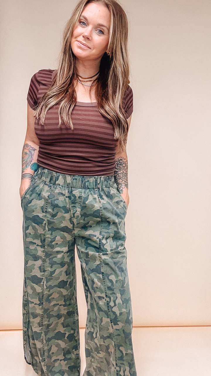 A woman modeling a brown striped short-sleeve top and green camouflage wide-leg pants against a plain beige studio background.