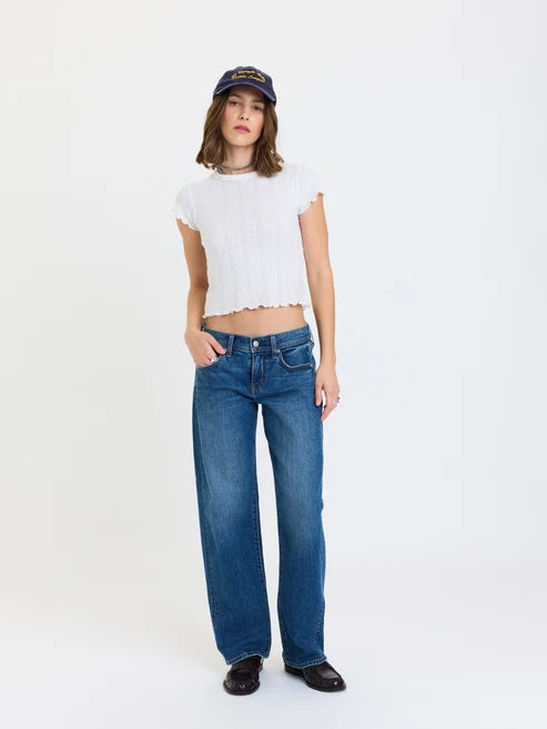 A model stands against a plain white studio background wearing a white cropped short-sleeve top, blue wide-leg jeans, black shoes, and a dark baseball cap.
