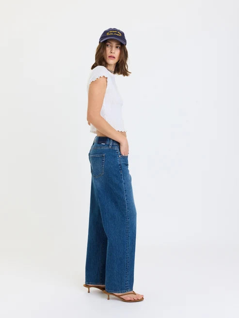 A woman modeling a pair of wide-leg blue denim jeans, paired with a white sleeveless top, a navy baseball cap, and brown heeled sandals, photographed against a solid white studio background.