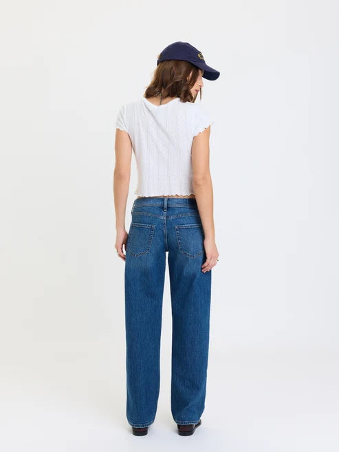 A model shown from the back wearing a white short-sleeve crop top, blue wide-leg jeans, and a navy blue baseball cap against a plain white studio background.