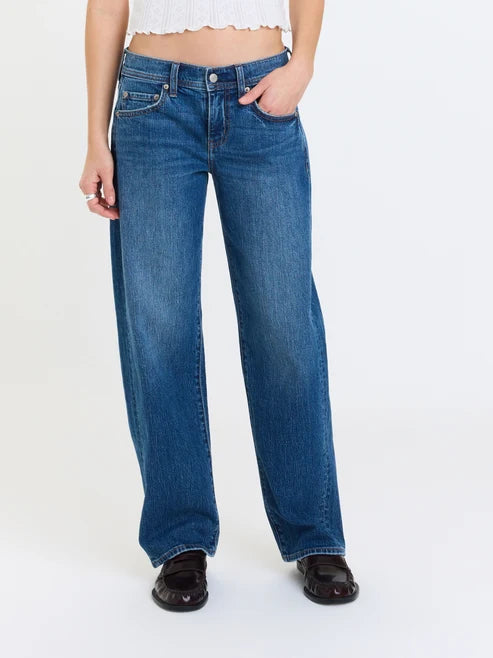 A person wearing blue wide-leg jeans with a white cropped top and black loafers, standing against a plain white studio background.