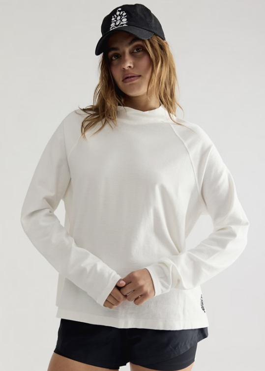 Woman wearing white long sleeve mock neck top, black shorts and black baseball cap on plain background