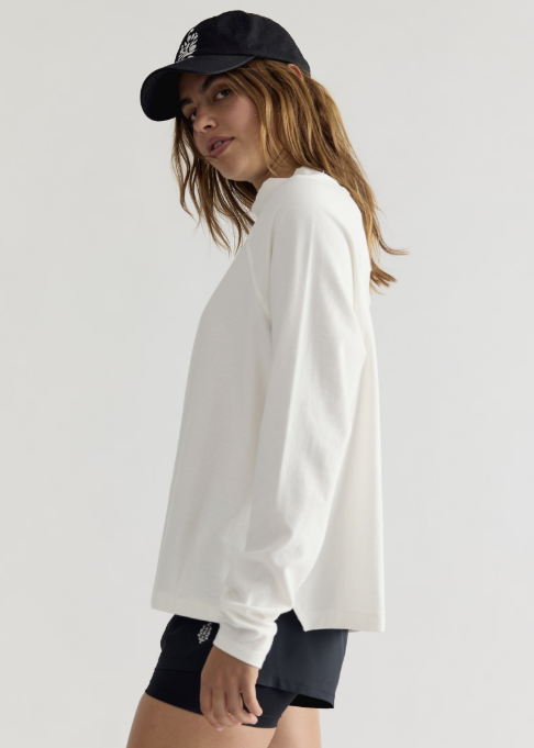 Woman wearing white long sleeve mock neck top, black shorts and black baseball cap on plain background
