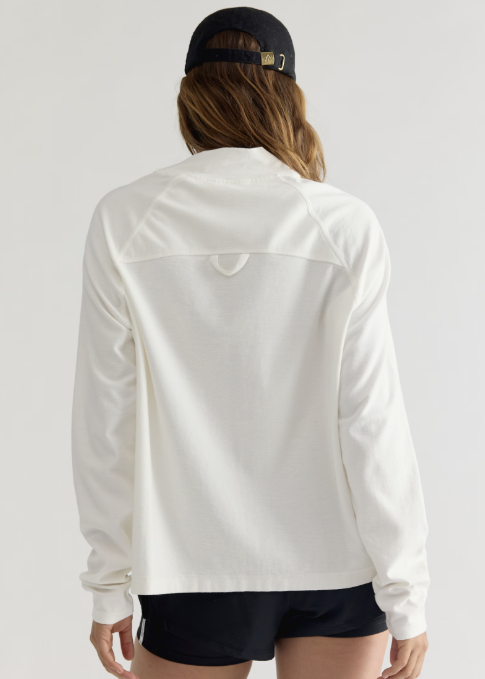 Woman shown from back wearing white long sleeve mock neck, black shorts and black baseball cap on plain background