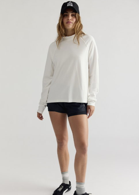 Woman wearing white long sleeve mock neck top, black shorts and black baseball cap on plain background