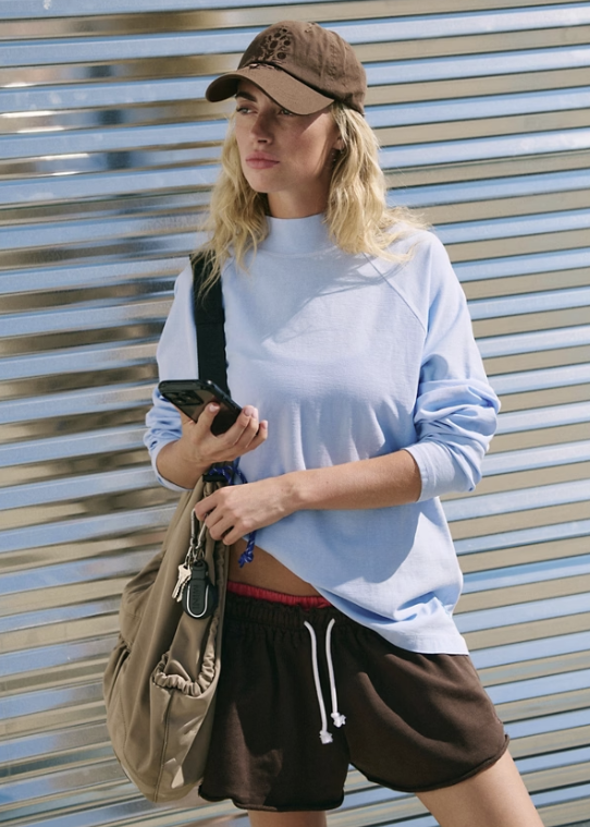 Woman wearing light blue long sleeve mock neck top, brown shorts and brown baseball cap outdoors