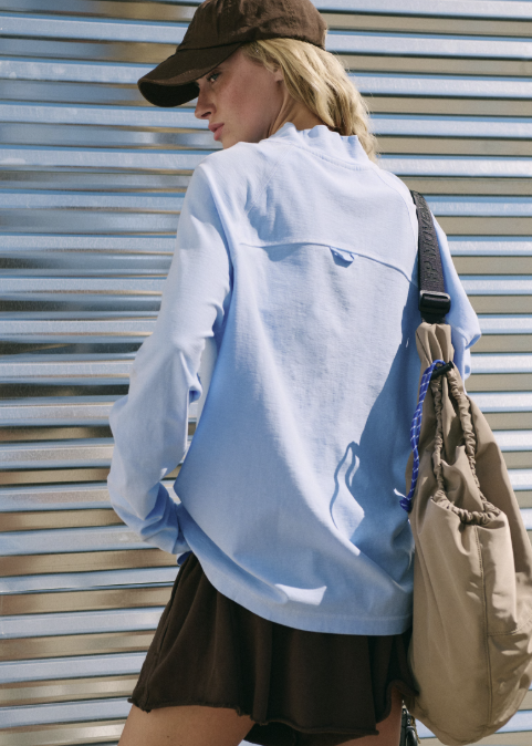 Woman shown from back wearing light blue long sleeve top, brown shorts and brown baseball cap standing outdoors.