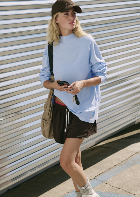 Woman wearing light blue long sleeve mock neck top, brown shorts and brown baseball cap standing outdoors.