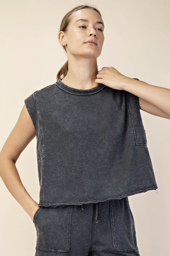 A woman wearing a dark gray washed sleeveless crop top with front patch pockets, paired with matching dark gray pants, against a solid beige background.