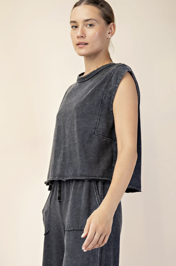 A woman wearing a dark gray washed sleeveless crop top and matching dark gray drawstring pants, photographed against a plain beige studio background.