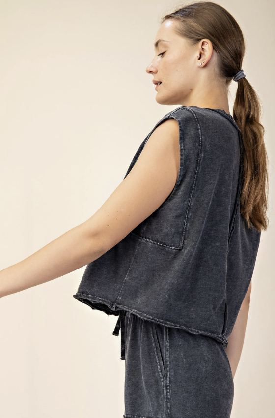 A woman shown from the side wearing a sleeveless dark gray washed denim vest and matching denim bottoms against a plain light beige studio background.