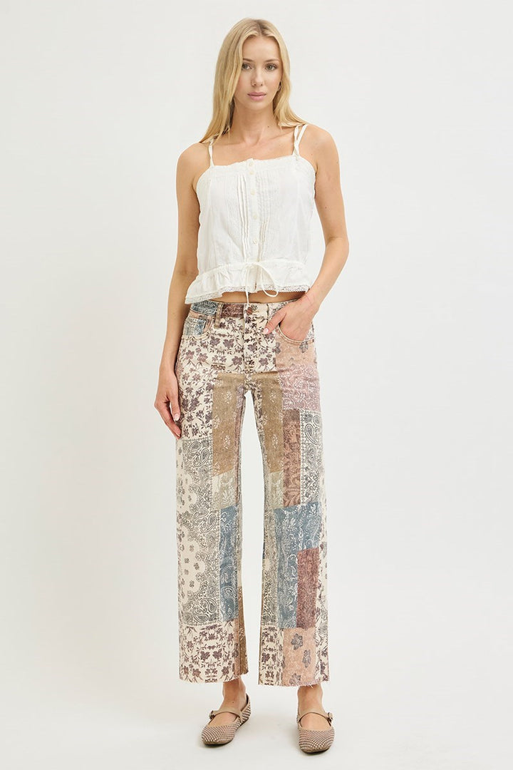 A woman modeling a white sleeveless top and high-waisted patchwork wide-leg pants in beige, brown, and muted blue tones, paired with beige flats, against a plain white studio background.