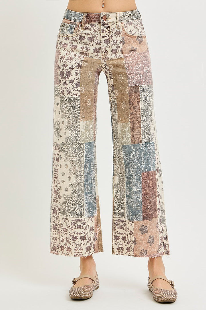A pair of wide-leg cropped pants featuring a multicolor patchwork print with floral and paisley patterns in beige, brown, blue, and pink tones, shown on a model wearing flat shoes against a light gray background.