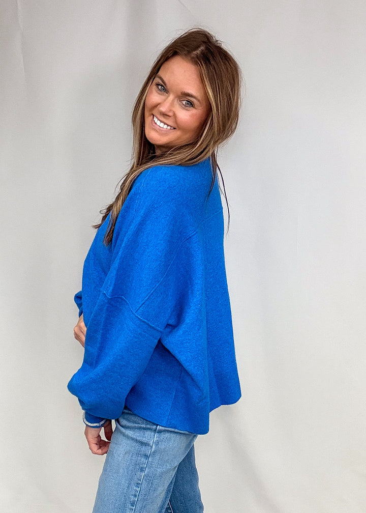 Woman wearing blue sweater with contrsasting trim on neck and sleeves with jeans on plain background