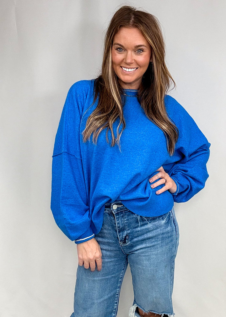 Woman wearing blue sweater with contrasting trim on neck and sleeves with jeans on plain background