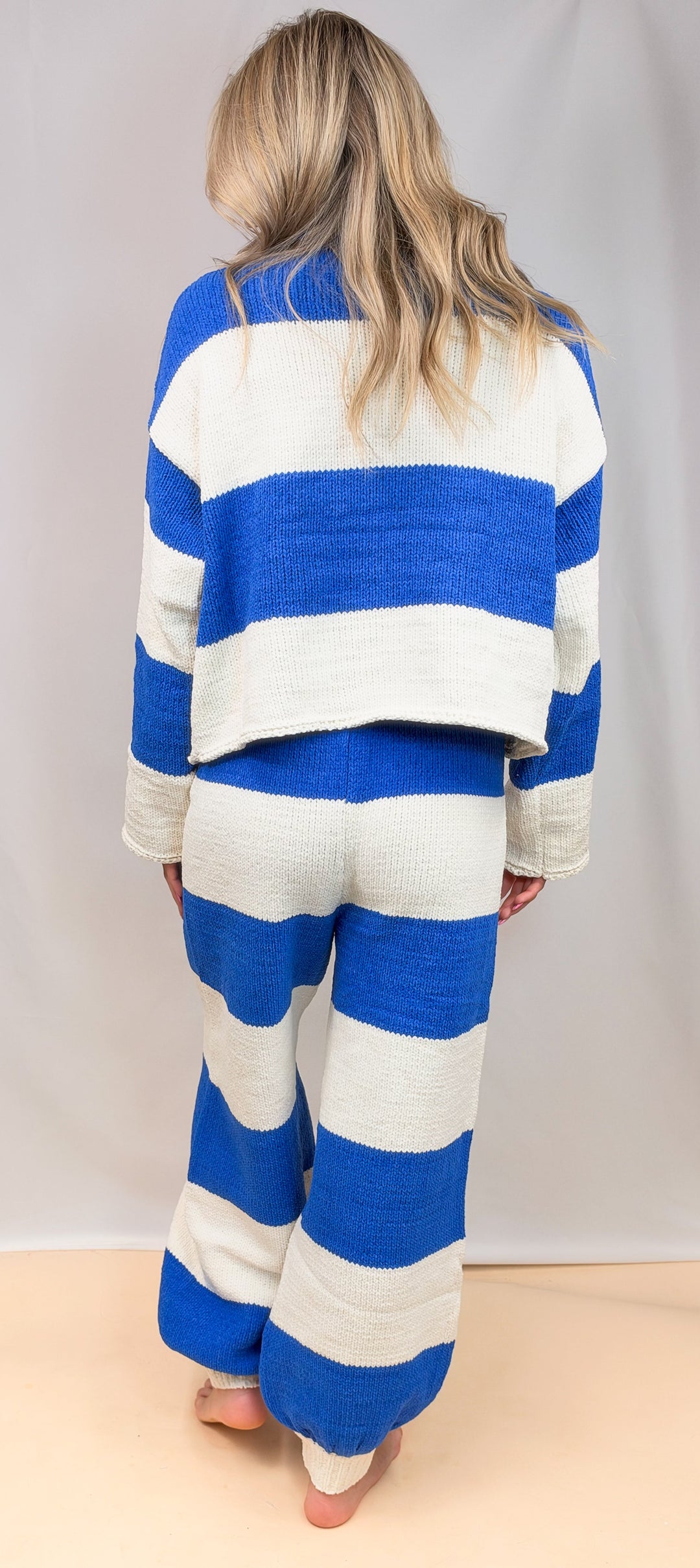A woman viewed from behind wearing a matching knit lounge set with bold blue and off-white horizontal stripes, standing against a plain light gray studio backdrop.