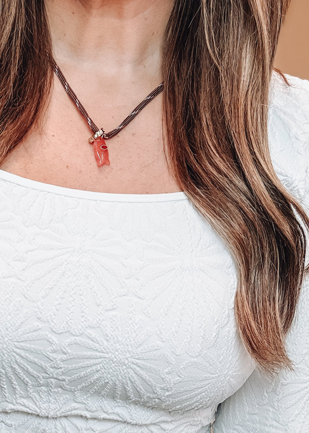 A close-up of a woman wearing a textured white long-sleeve top and a brown cord necklace with a small red pendant, paired with blue jeans.