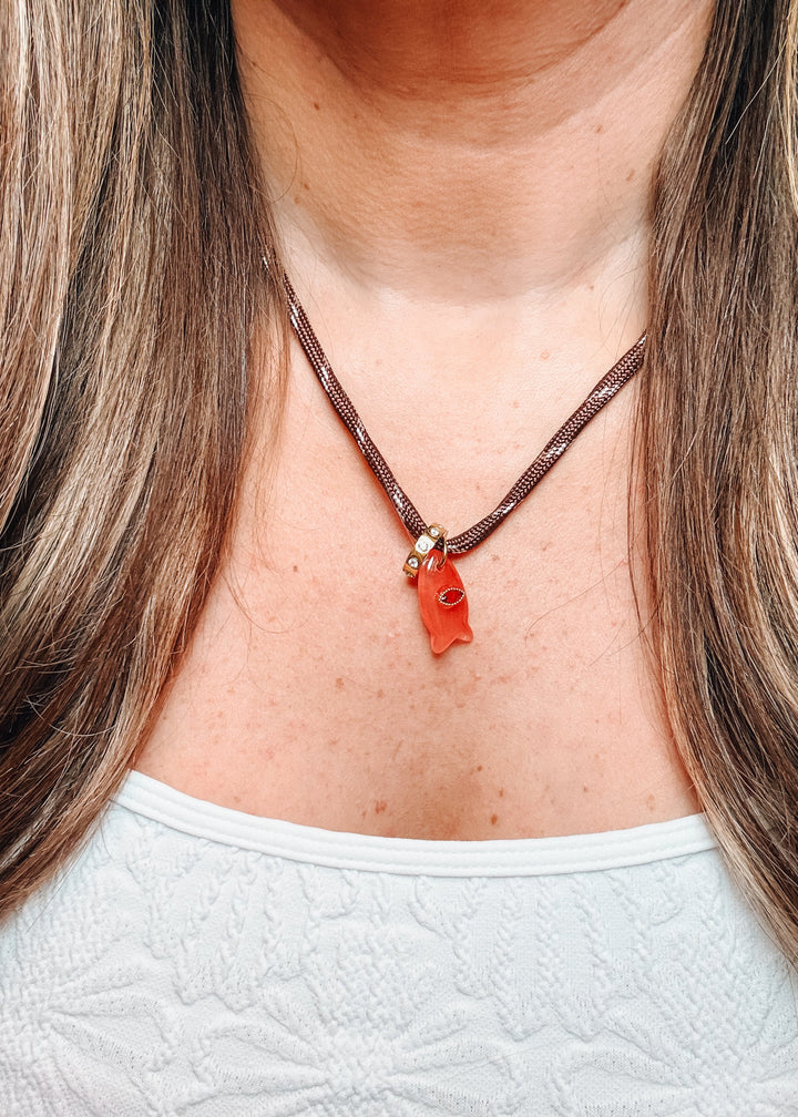A close-up of a person wearing a brown braided cord necklace with a small orange pendant and gold-toned beads.