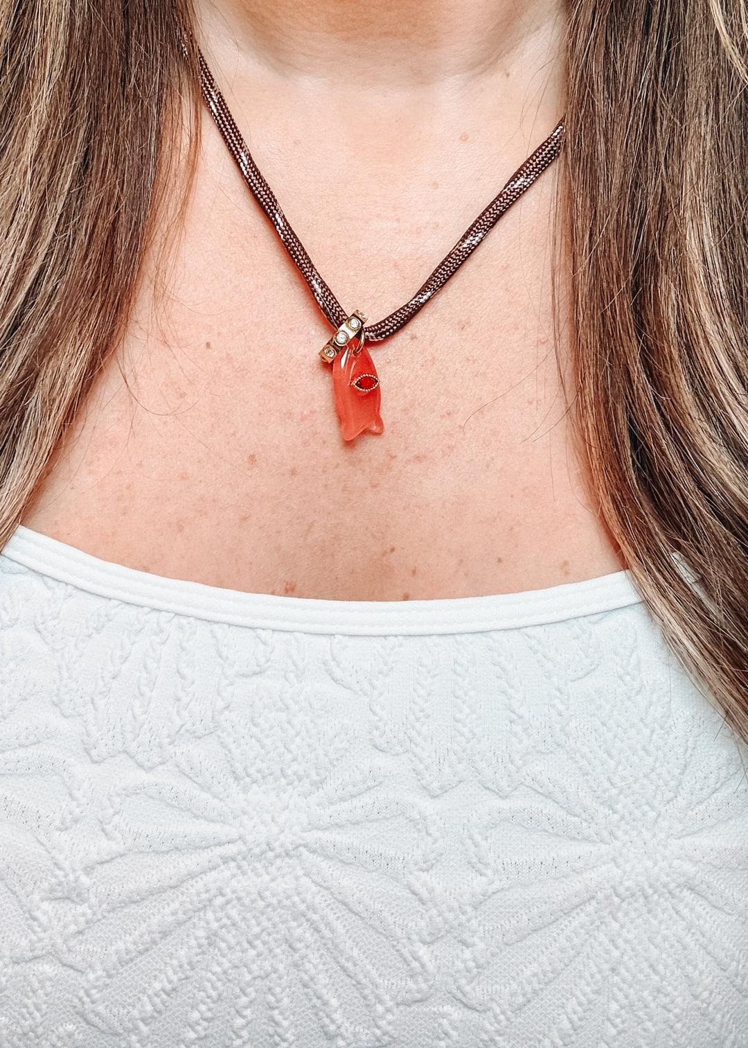 A close-up of a woman wearing a brown braided cord necklace with a small carved orange-red pendant, paired with a white textured top.