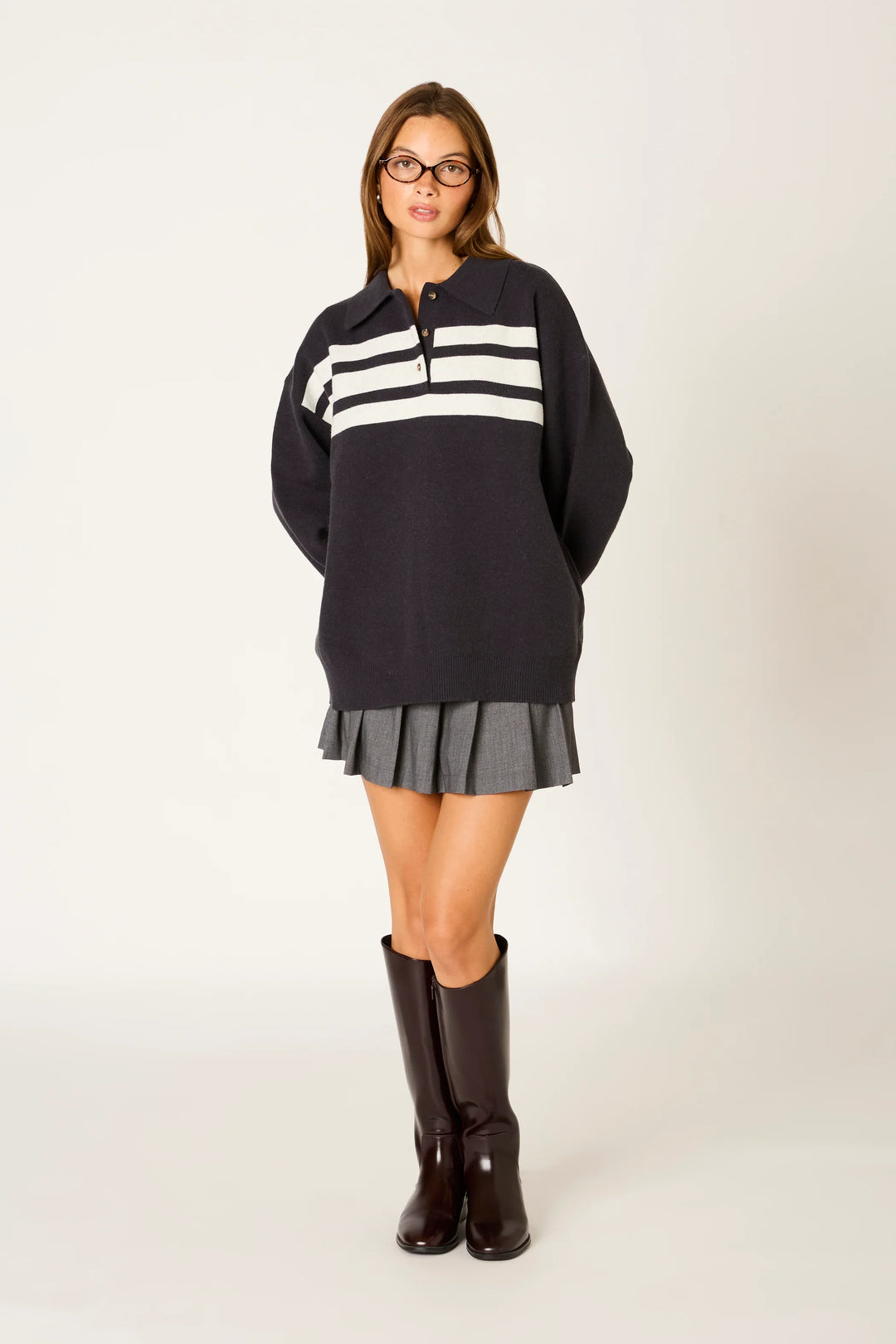 A woman modeling a dark navy long-sleeve sweater with three horizontal white stripes across the chest, paired with a gray pleated mini skirt and dark brown knee-high boots, against a plain light background.