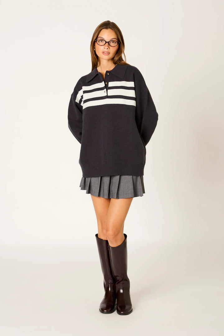 A woman modeling a dark navy long-sleeve sweater with three horizontal white stripes across the chest, paired with a gray pleated mini skirt and dark brown knee-high boots, against a plain light background.