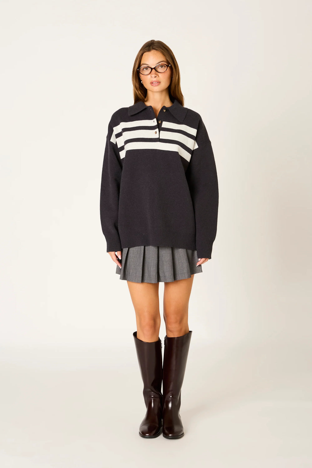 A woman modeling a dark navy long-sleeve polo sweater dress with white horizontal stripes across the chest, paired with a gray pleated mini skirt and dark brown knee-high boots, against a plain light gray studio background.