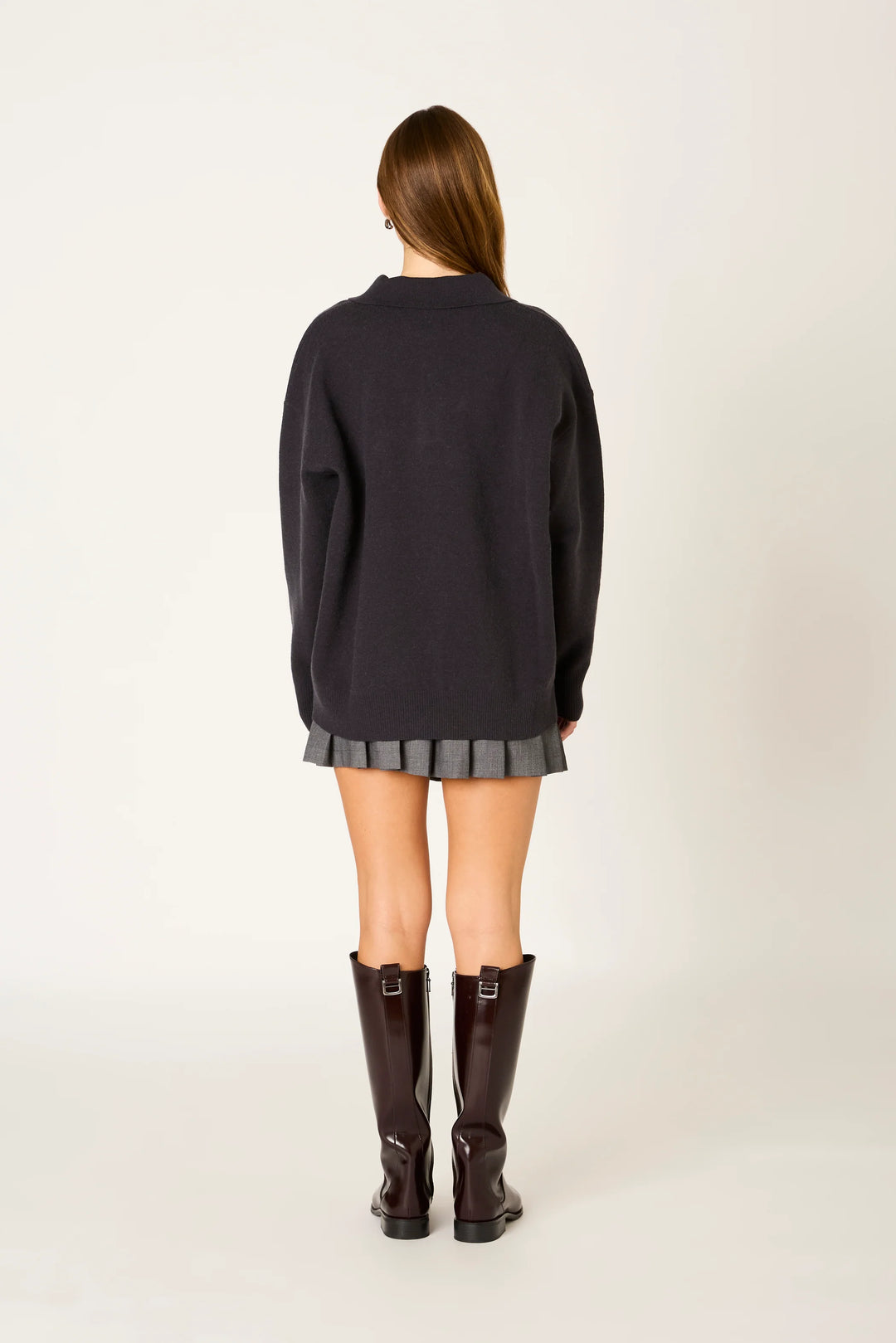 A woman viewed from the back wearing an oversized dark charcoal knit sweater, a gray pleated mini skirt, and dark brown knee-high boots against a plain off-white studio background.