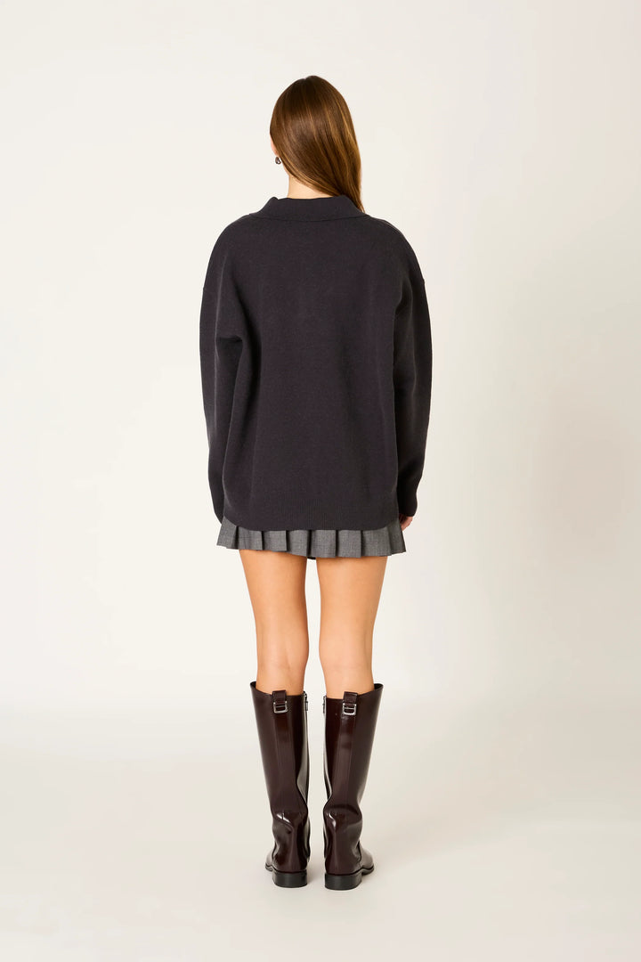 A woman viewed from the back wearing an oversized dark charcoal knit sweater, a gray pleated mini skirt, and dark brown knee-high boots against a plain off-white studio background.