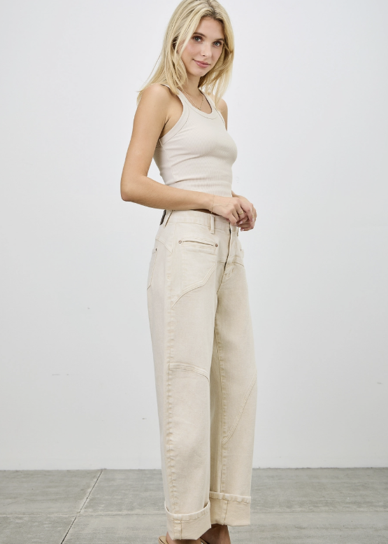 A blonde model wearing a beige ribbed tank top and matching beige wide-leg cropped jeans with rolled cuffs, paired with clear strap heels, standing against a light gray studio wall.