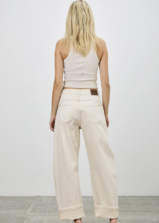 A woman viewed from behind wearing off-white wide-leg jeans with rolled cuffs, a ribbed white tank top, and beige high heels against a plain white wall and gray floor.