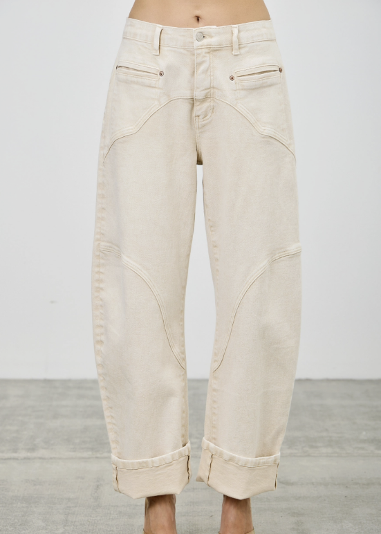 A person from the waist down wearing off-white or cream-colored wide-leg cropped jeans with cuffed hems and metallic gold high-heeled sandals, standing against a light gray wall and concrete floor.