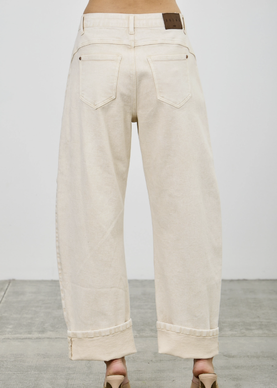 Back view of a person wearing off-white cropped wide-leg jeans with rolled cuffs and nude high-heeled sandals, standing against a light gray wall and concrete floor.