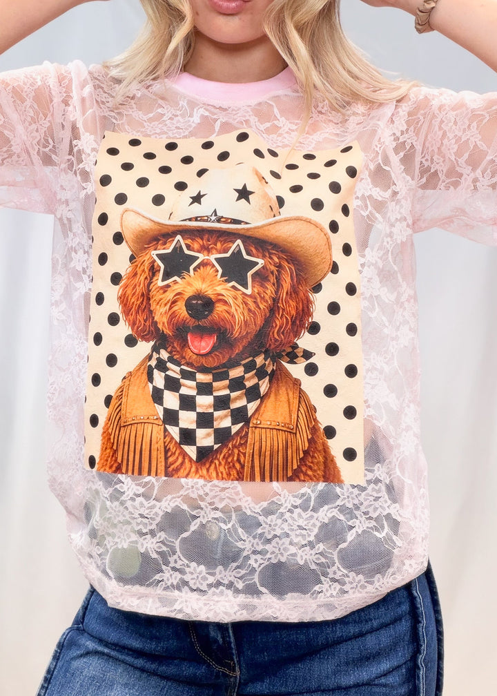 Woman wearing pink lace tee with poodle western graphic on front with denim jeans on plain background