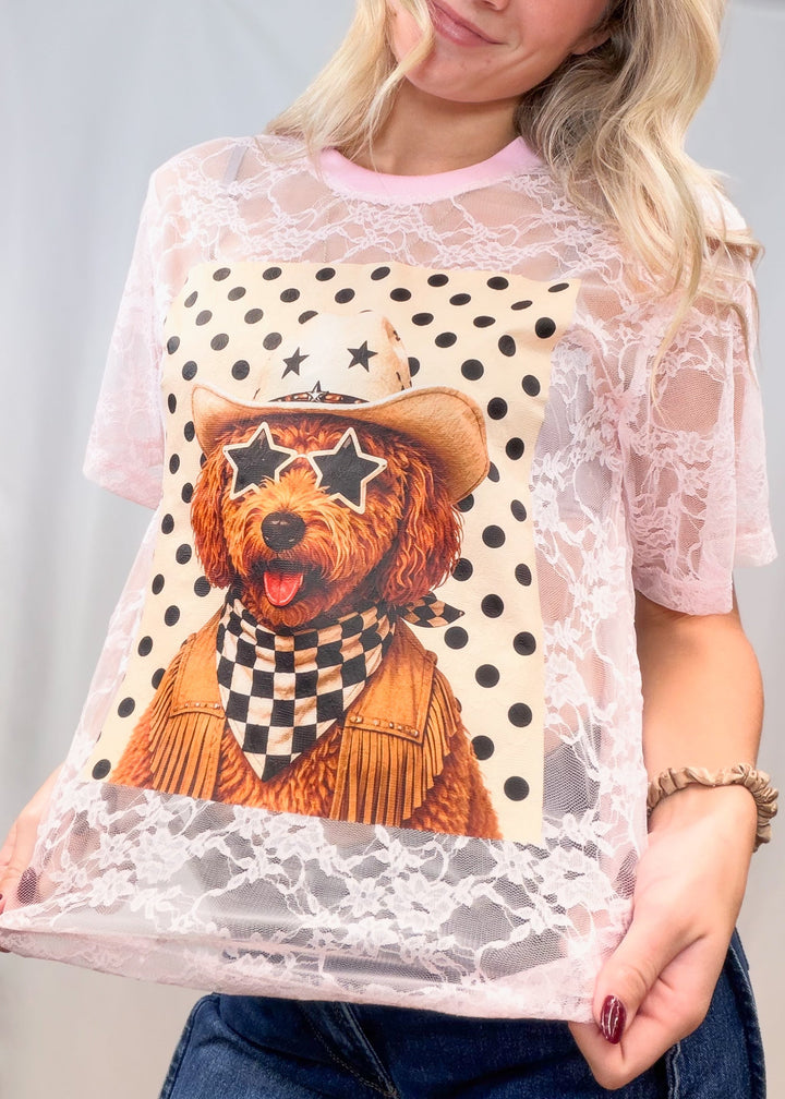 Woman wearing pink lace tee with poodle western graphic on front with denim jeans on plain background