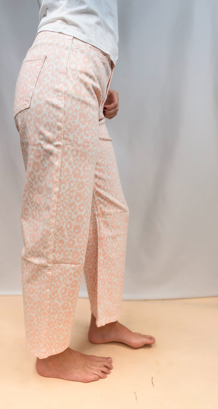 Woman shown from the side waist down wearing white tee and pink leopard pants on plain background