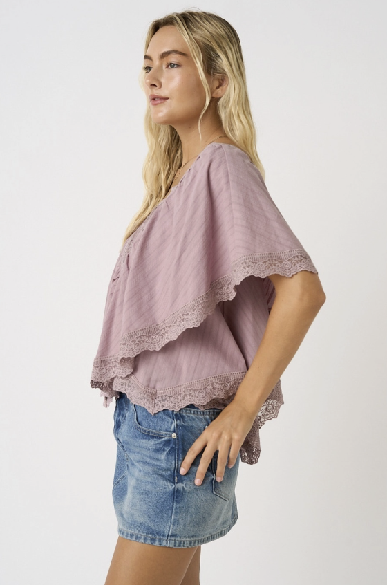 A woman wearing a dusty mauve short-sleeve top with layered ruffles and lace trim, paired with a denim mini skirt, photographed against a plain light gray studio background.