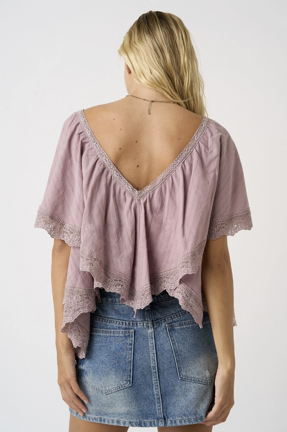 A woman shown from the back wearing a dusty mauve short-sleeve top with a deep V-neck and lace trim, paired with a blue denim skirt, against a plain white background.