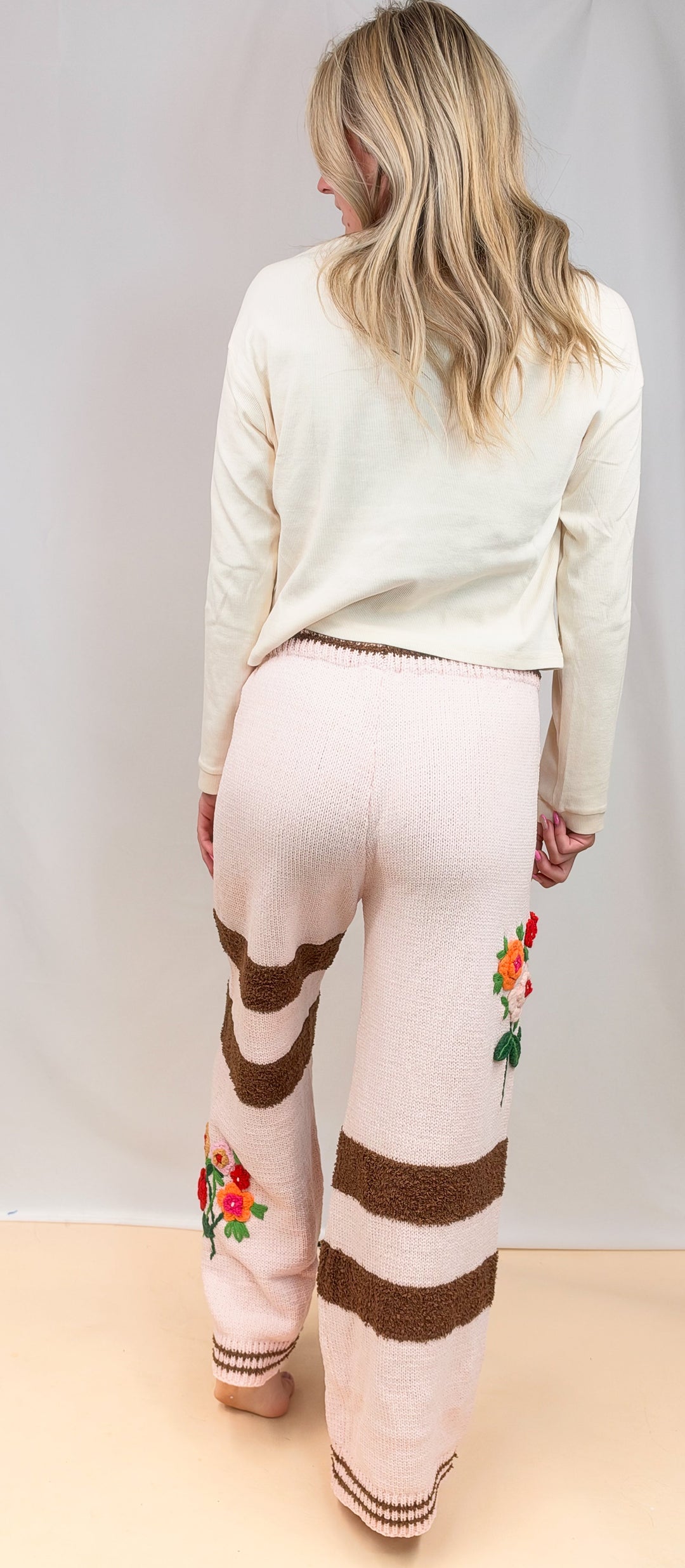 A woman viewed from behind wearing cream knit pants with brown horizontal stripes and colorful floral embroidery near the lower legs, paired with a cream long-sleeve top against a plain studio backdrop.