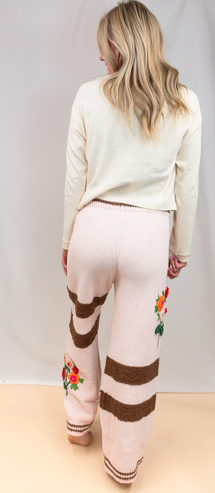 A woman viewed from behind wearing cream knit pants with brown horizontal stripes and colorful floral embroidery near the lower legs, paired with a cream long-sleeve top against a plain studio backdrop.