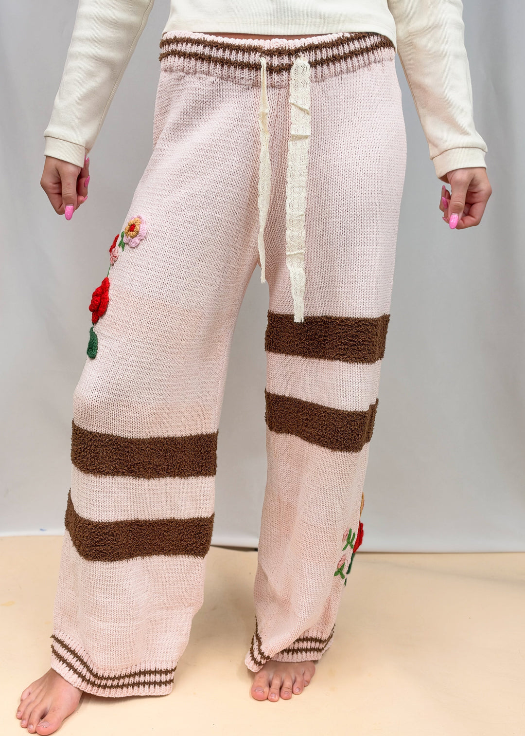 A person wearing loose, wide-leg knit pants in a pale blush pink and brown striped pattern, with small embroidered floral details on the legs. The photo is cropped from the chest down against a plain light gray studio backdrop.