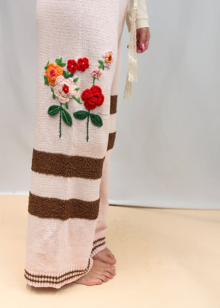 Woman shown from side view wearing pink knit pants with crochet flowers and brown stripes on plain background
