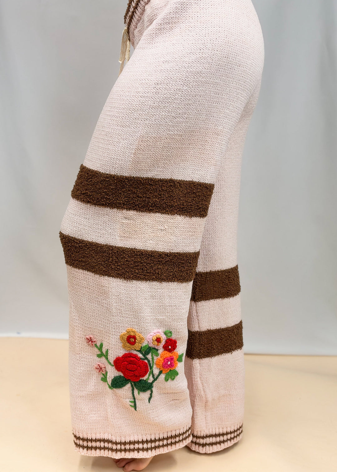 A close-up side view of a person wearing wide-leg knit pants in a light blush pink color with bold brown horizontal stripes and colorful floral embroidery near the lower left leg.
