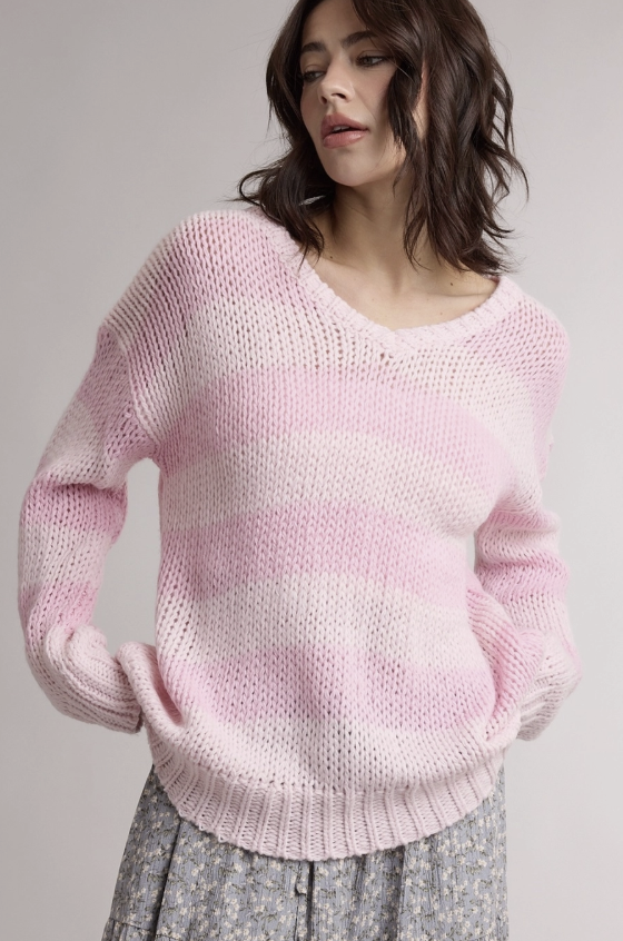 A woman wearing a chunky knit long-sleeve sweater in a soft pink and white horizontal stripe pattern, paired with a gray patterned skirt, against a plain light gray background.
