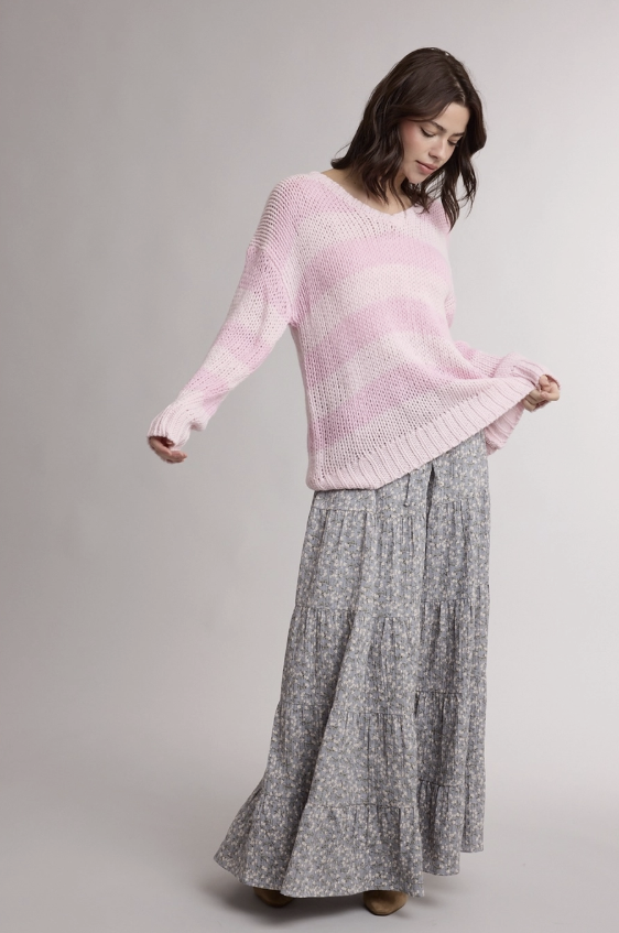 A woman modeling a light pink knit sweater paired with a long gray patterned skirt, photographed against a neutral light gray studio background.