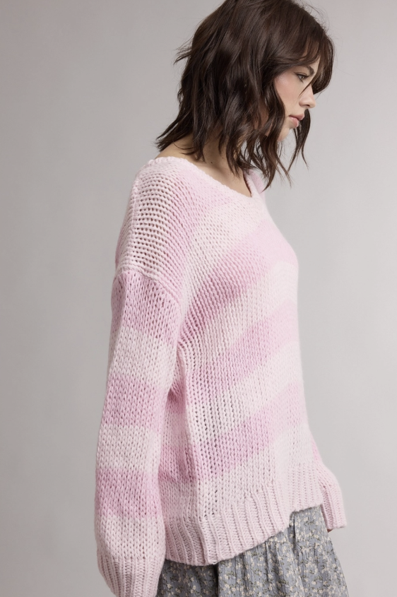 A woman wearing a light pink chunky knit sweater with horizontal darker pink stripes, paired with a gray patterned skirt, photographed against a neutral light gray background.