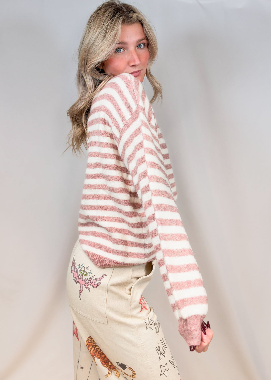 Woman shown from side view wearing pink and cream stripe mock neck sweater with beige pants with doodles on plain background