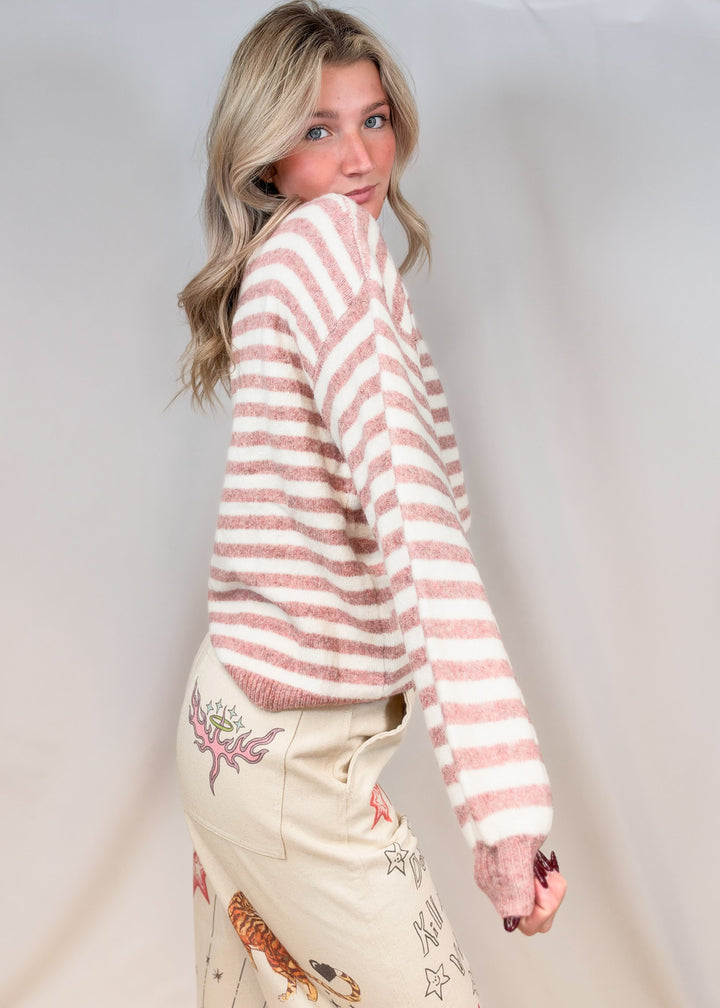 Woman shown from side view wearing pink and cream stripe mock neck sweater with beige pants with doodles on plain background