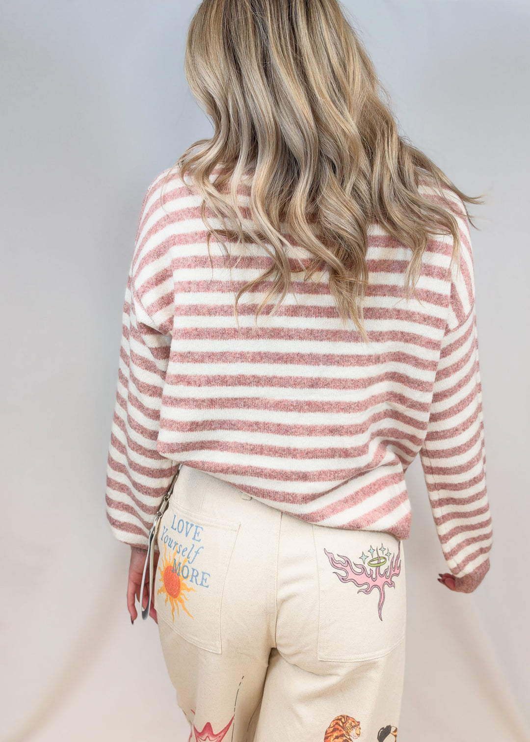 Woman shown from back view wearing pink and cream stripe sweater with beige pants on plain background