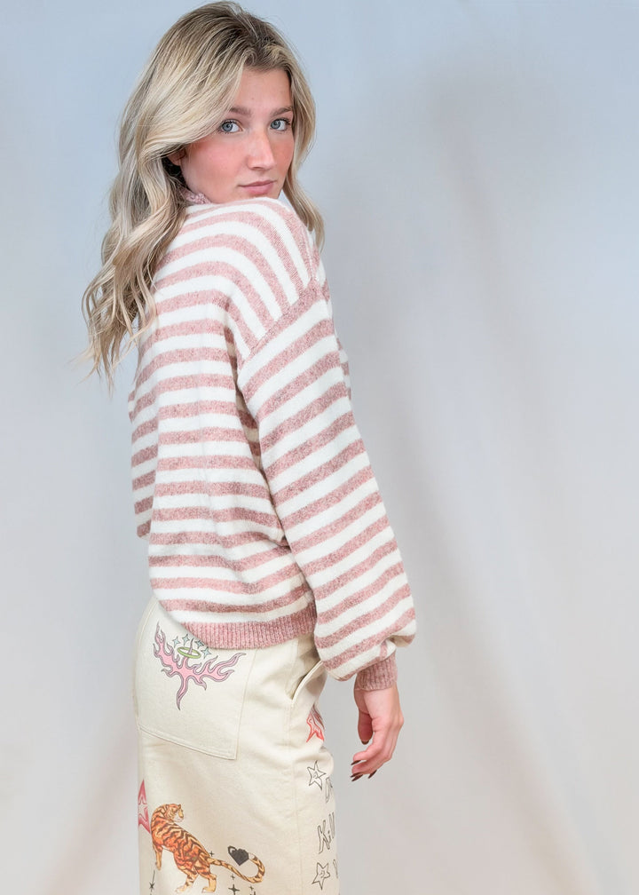 Woman shown from side view wearing pink and cream sweater with beige doodle pants on plain background